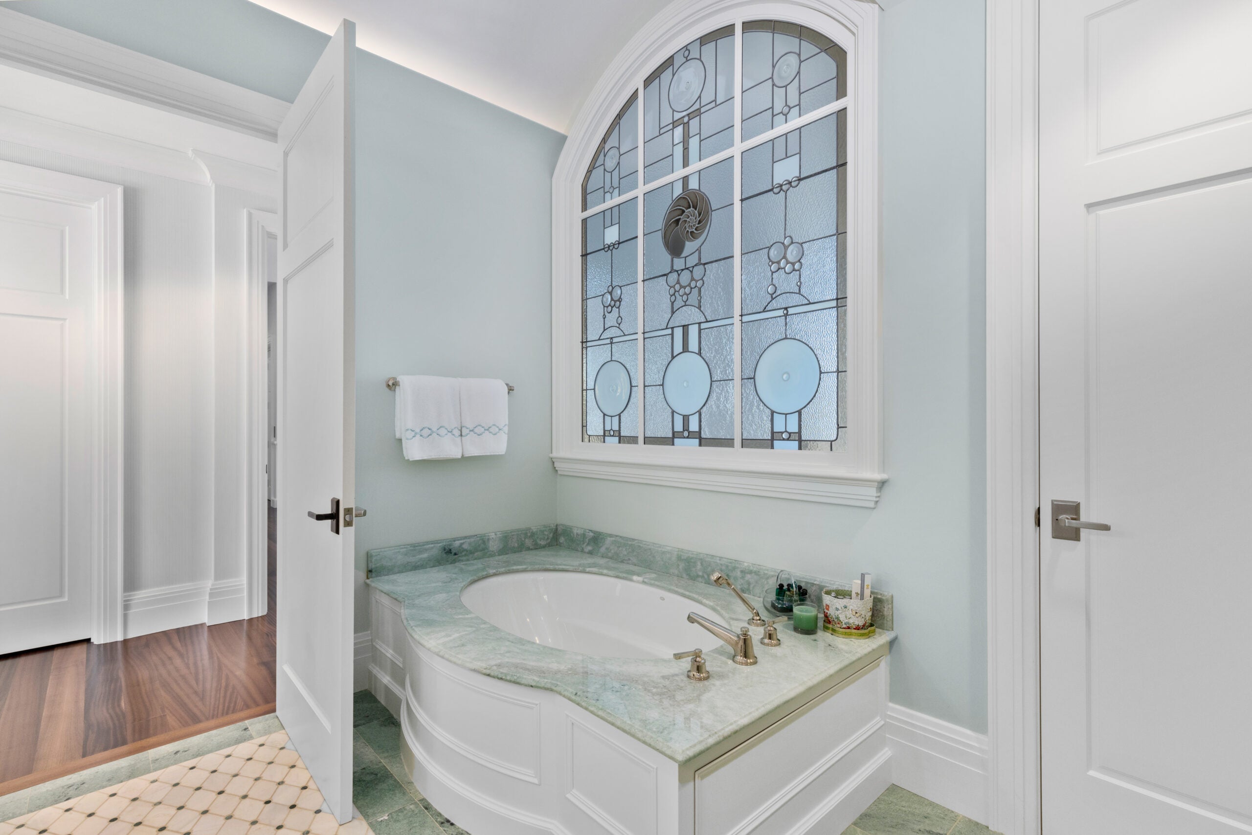 A door is open to a bathroom with a green marble tub and floor. The tub, which has gold fixtures, is oval and set before a stained-glass window depicting bubbles and a seashell.