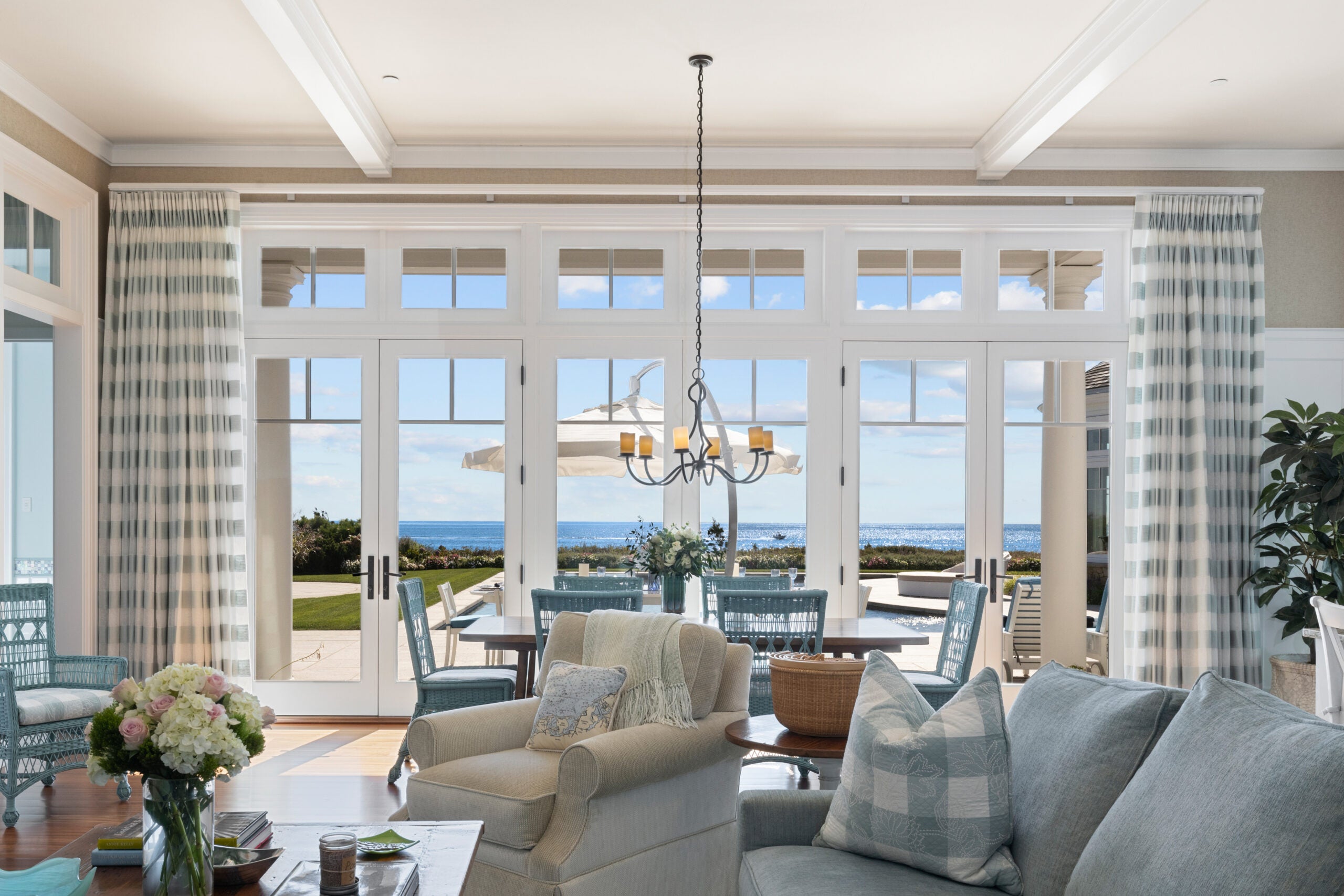 A six-seat dining area with light blue whicker chairs before a trio of glass doors. The view is of a sunny day and the ocean. The living room has stuffed chairs of gray and heather blue. The curtains and one of the throw pillows on the couch are white-and-blue buffalo check.