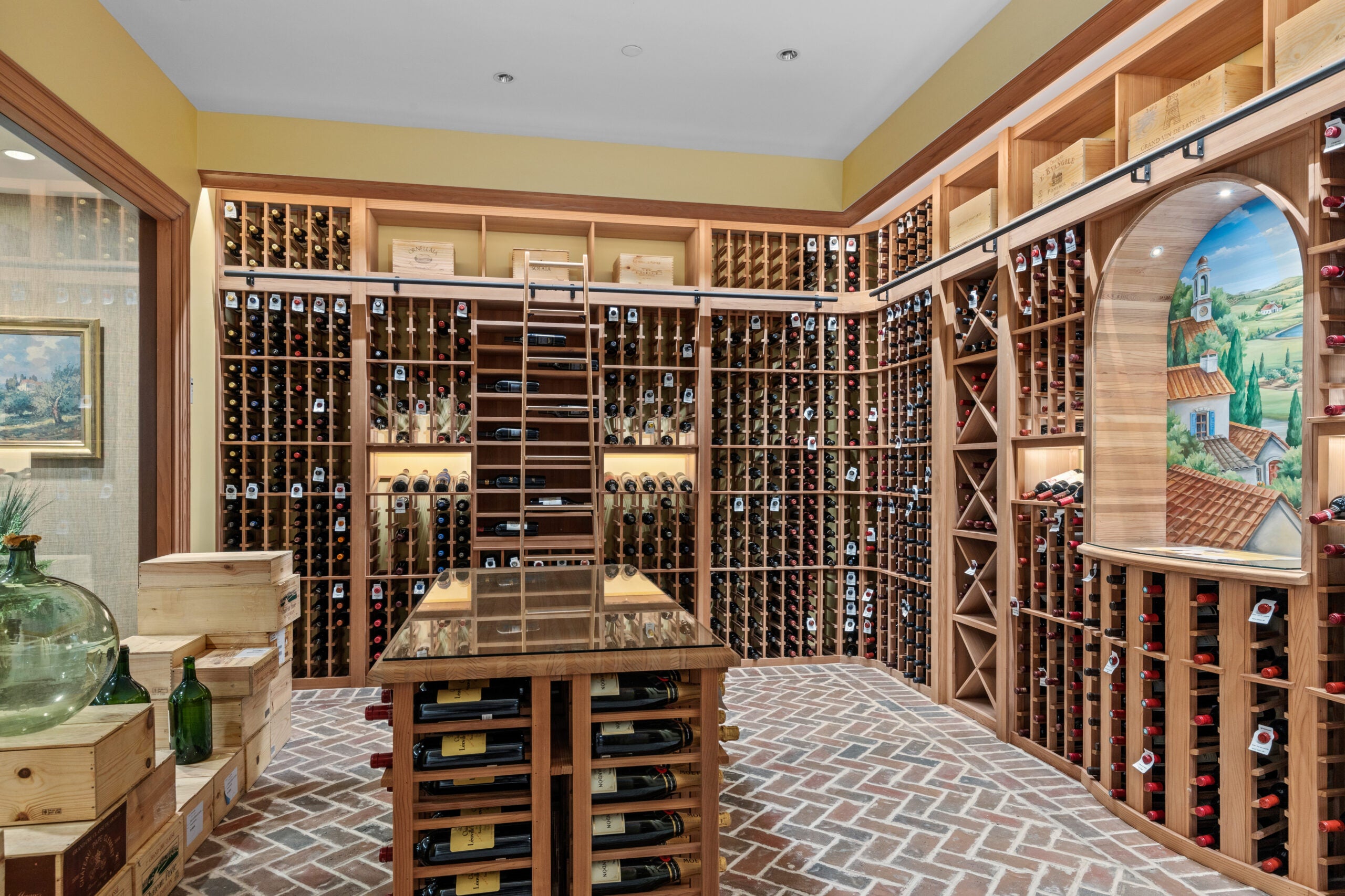 A view of a multi-bottle wine cellar with a brick floor laid in a herringbone pattern. There are several wine crates stacked off to the left. The door to the cellar is glass.