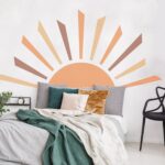 A decal of an orange sun with brown and orange rays that serves as a headboard. The bed has white and green bedding and gray, green, gold, and white throw pillows. There is a small open nightstand and an open backed chair in the room as well.