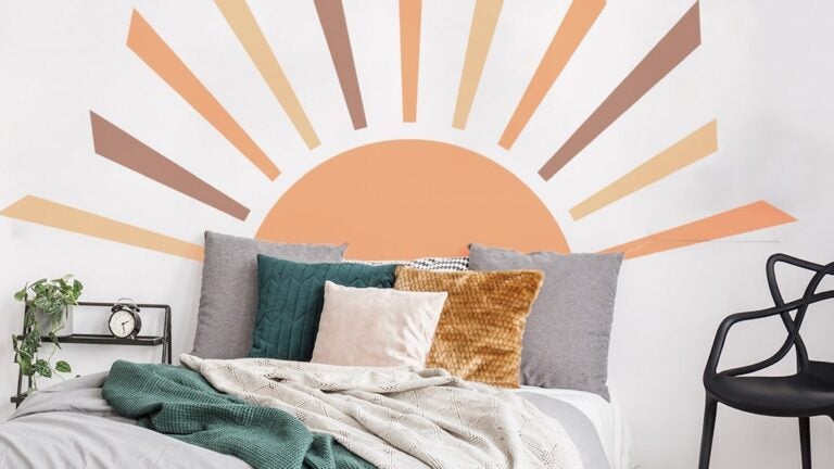 A decal of an orange sun with brown and orange rays that serves as a headboard. The bed has white and green bedding and gray, green, gold, and white throw pillows. There is a small open nightstand and an open backed chair in the room as well.