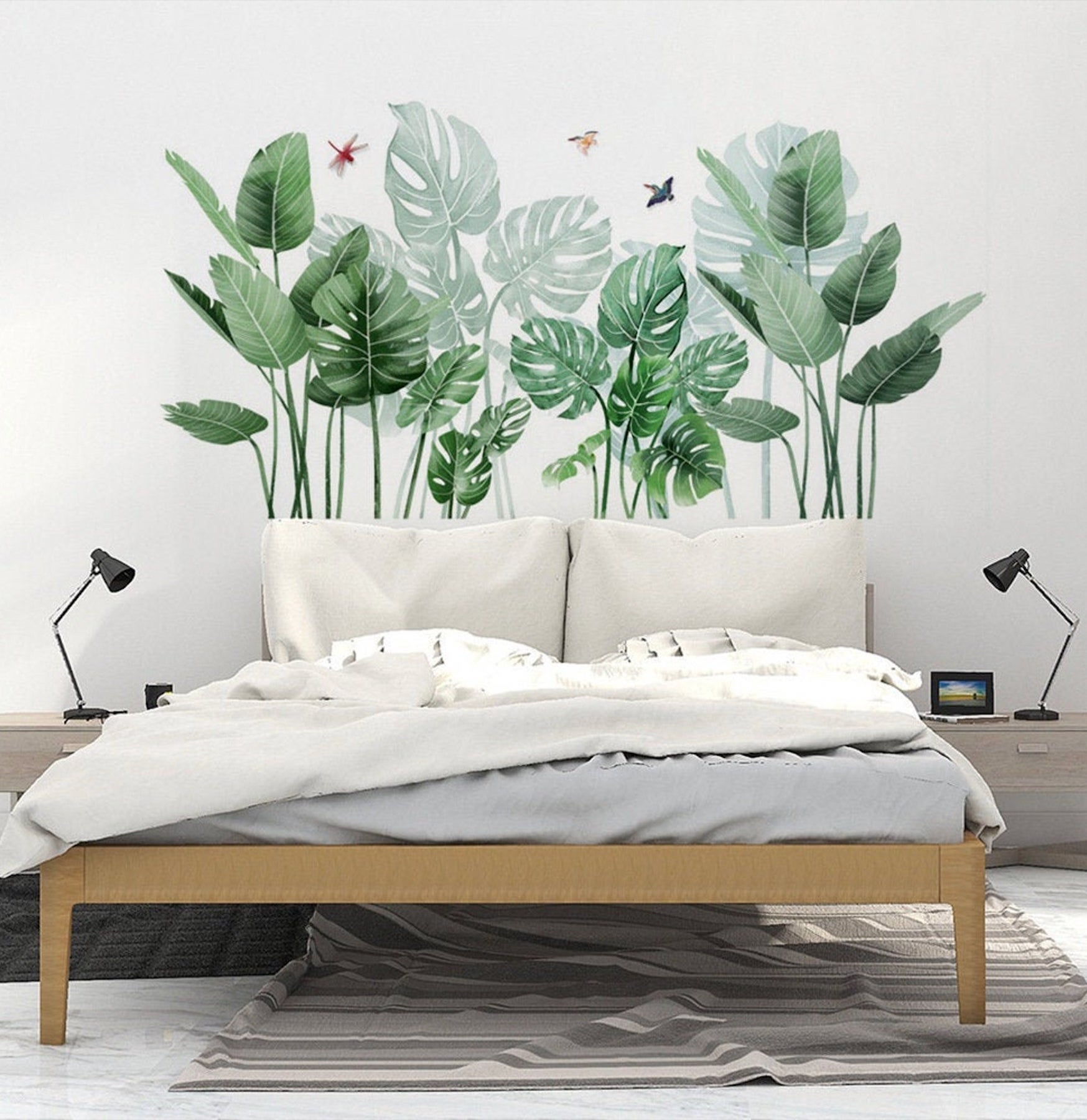 A tall decal of a monstera leaf with butterflies serves as a headboard for a wooden, skinny-legged bed with white and gray bedding. End tables the color of driftwood with desk lamps flank the bed.