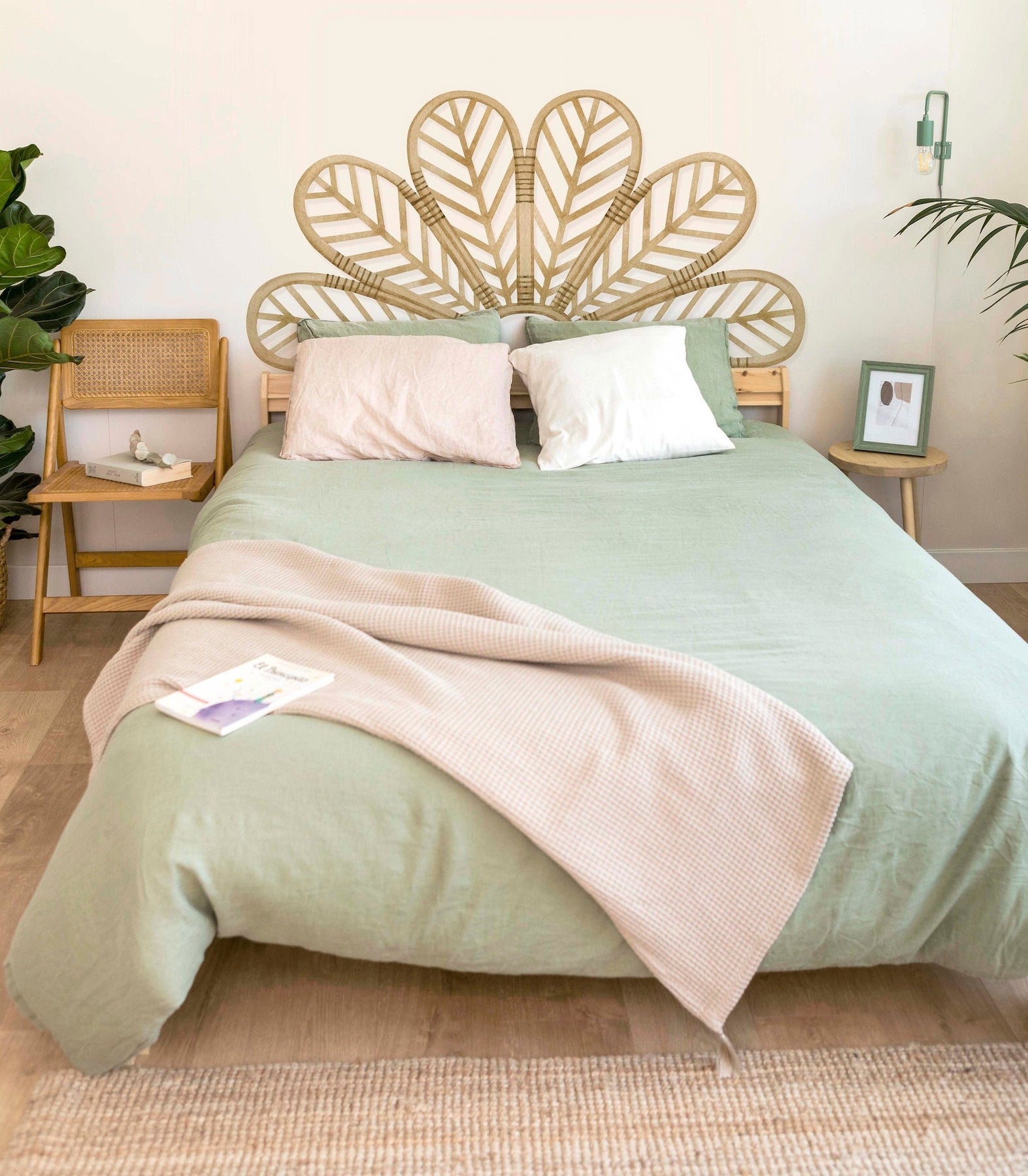 A decal that looks like wicker peacock feathers serves as a headboard for a bed with a green comforter and white and pink pillows. There is a folding chair in the background and a monstera leaf plant.