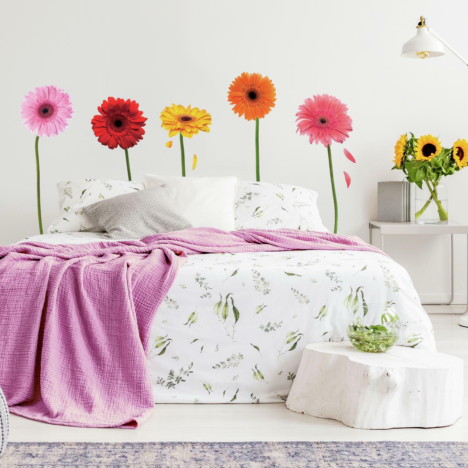 A decale with tall-stemmed Gerber daisies in pink red, yellow, and orange serves as a headboard for a bed with a pink blanket and floral bedding. An end table the color of driftwood holds a vase filled with sunflowers.
