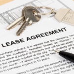 A document with the words "LEASE AGREEMENT" written in ALLCAPS. A set of keys with a house key fob rests just above it, on top of a floor plan. A black ballpoint pen rests on the lease document, pointing toward the keys.