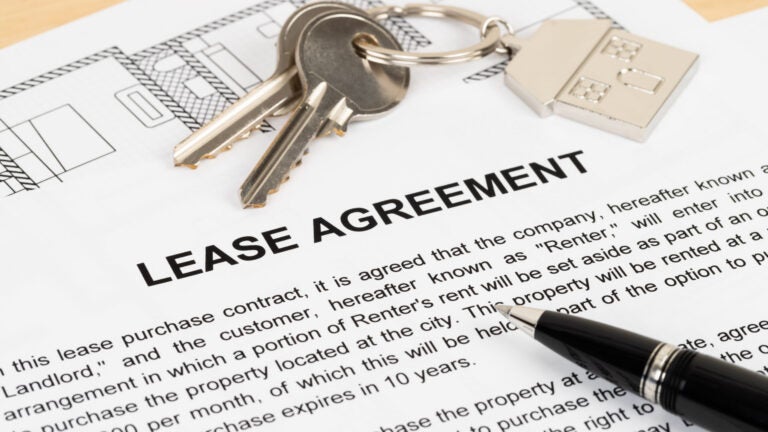 A document with the words "LEASE AGREEMENT" written in ALLCAPS. A set of keys with a house key fob rests just above it, on top of a floor plan. A black ballpoint pen rests on the lease document, pointing toward the keys.