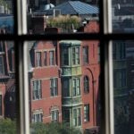 A view of brick multi-story townhomes with copper oriel windows. Leafy trees are in the bottom left corner of the photo. The view is as seen through a multi-paned window.
