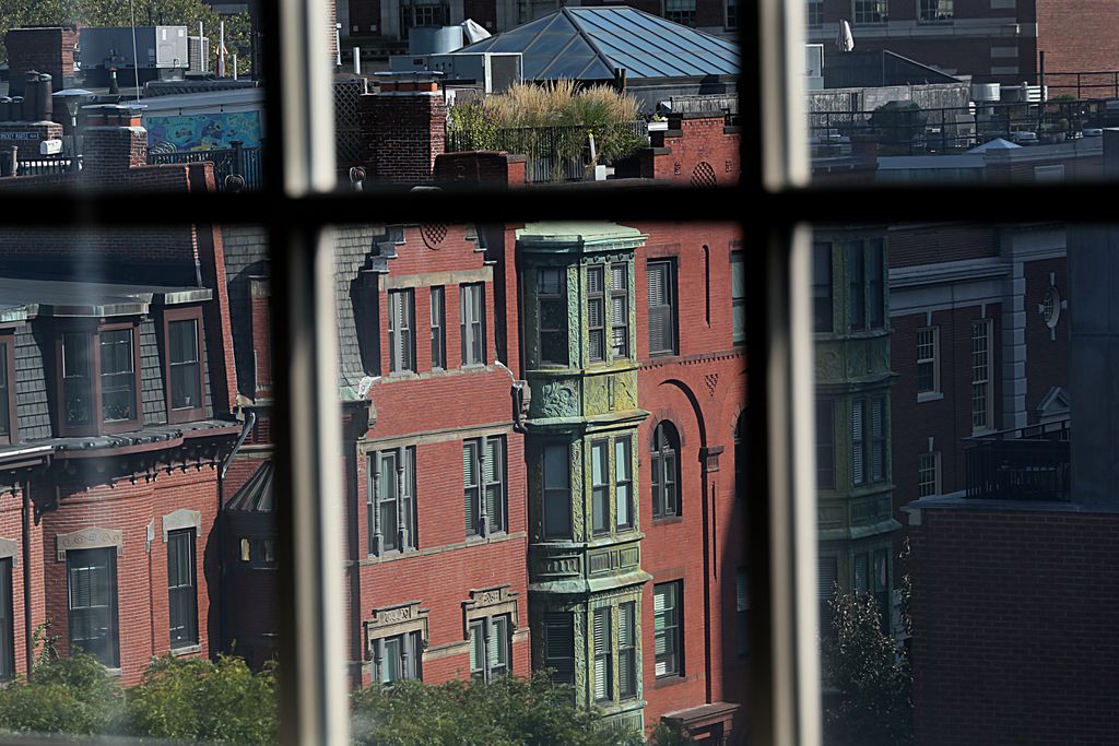 A view of brick multi-story townhomes with copper oriel windows. Leafy trees are in the bottom left corner of the photo. The view is as seen through a multi-paned window.