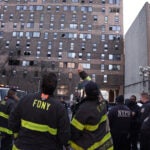 Firefighters stand outside a Bronx brick high-rise that was consumed by fire. Several windows in the building are broken.