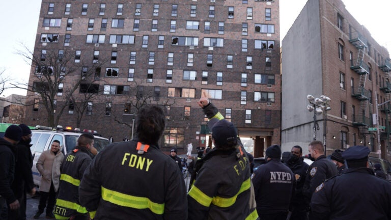 Firefighters stand outside a Bronx brick high-rise that was consumed by fire. Several windows in the building are broken.
