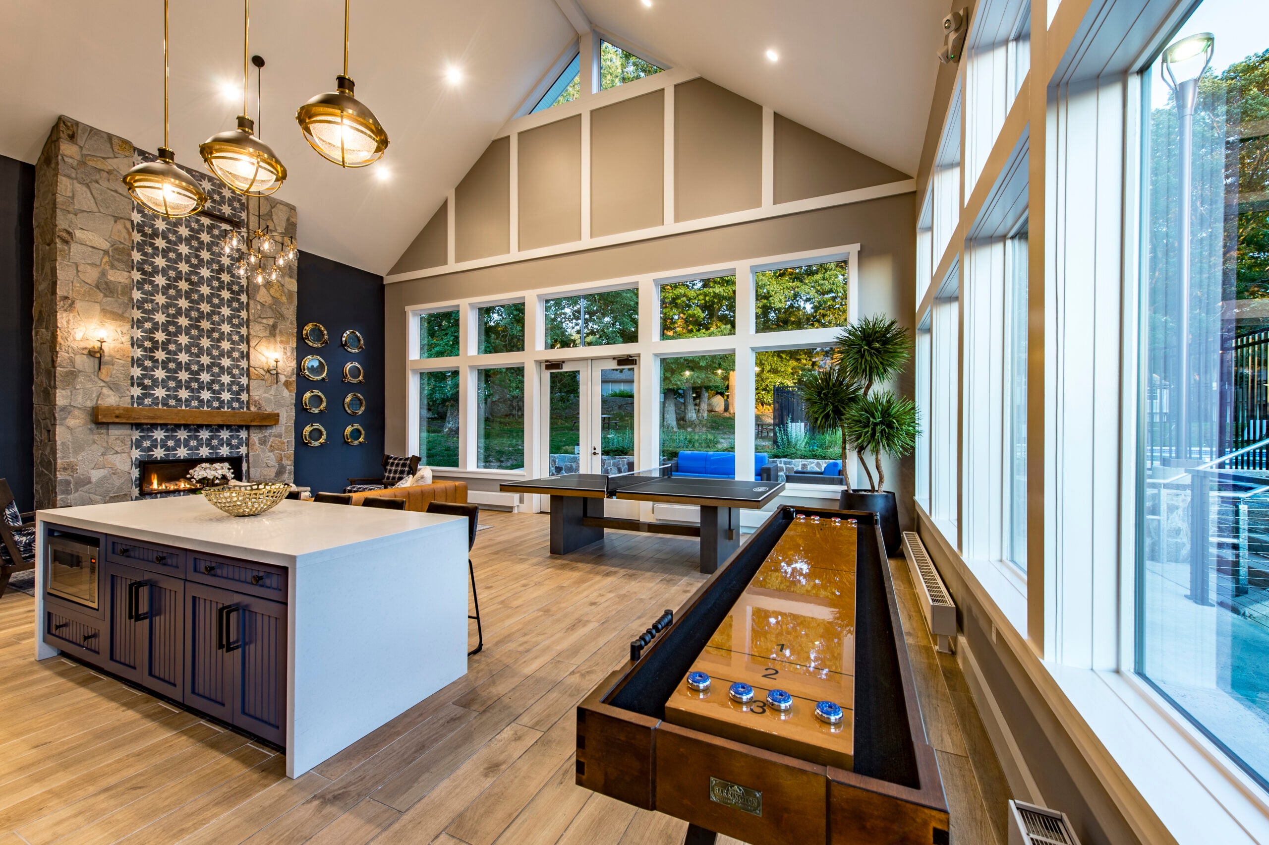 A two-story room with a vaulted ceiling, several industrial lights hanging from the ceiling, game tables, a fireplace, and a counter with a waterfall edge and seating. The color scheme is beige, white, and black. Expanses of windows line the walls. The flooring looks like wood.