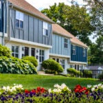 The exterior of several attached town homes, which feature white lower floors and upper floors with vertical wood, Stick-style-like siding. The town homes are navy, gray, and a grayish green. Bush line the front of the homes. There is an expanse of lawn with red and white flowers. Mature trees are full of leaves in the background. The sky is overcast.