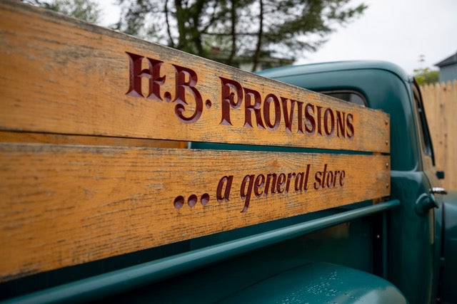 A antique green truck with a bed with wooden walls. H.B. Provisions is etched into the wood.