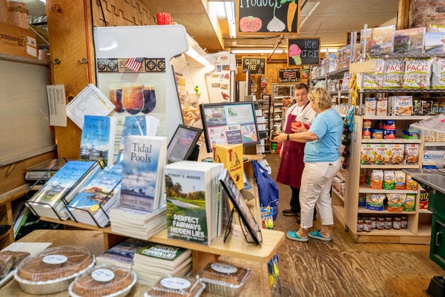 Baked goods and books about golf and tidal pools line a table in the foreground as a customer and an employee talk amid the shelves in the background.