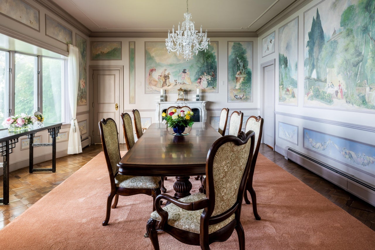 The walls of the formal dining room are decorated with soft-colored murals. There is a long dinner table in the center of the room under a glass chandelier. There are seven chairs set up around the table, which is sitting on a light rose colored rug.