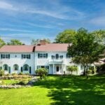 The exterior of the colonial-style home is white with a red-brown roof. There is a large, green yard in front of the home, and a water fountain to the left of the entrance.