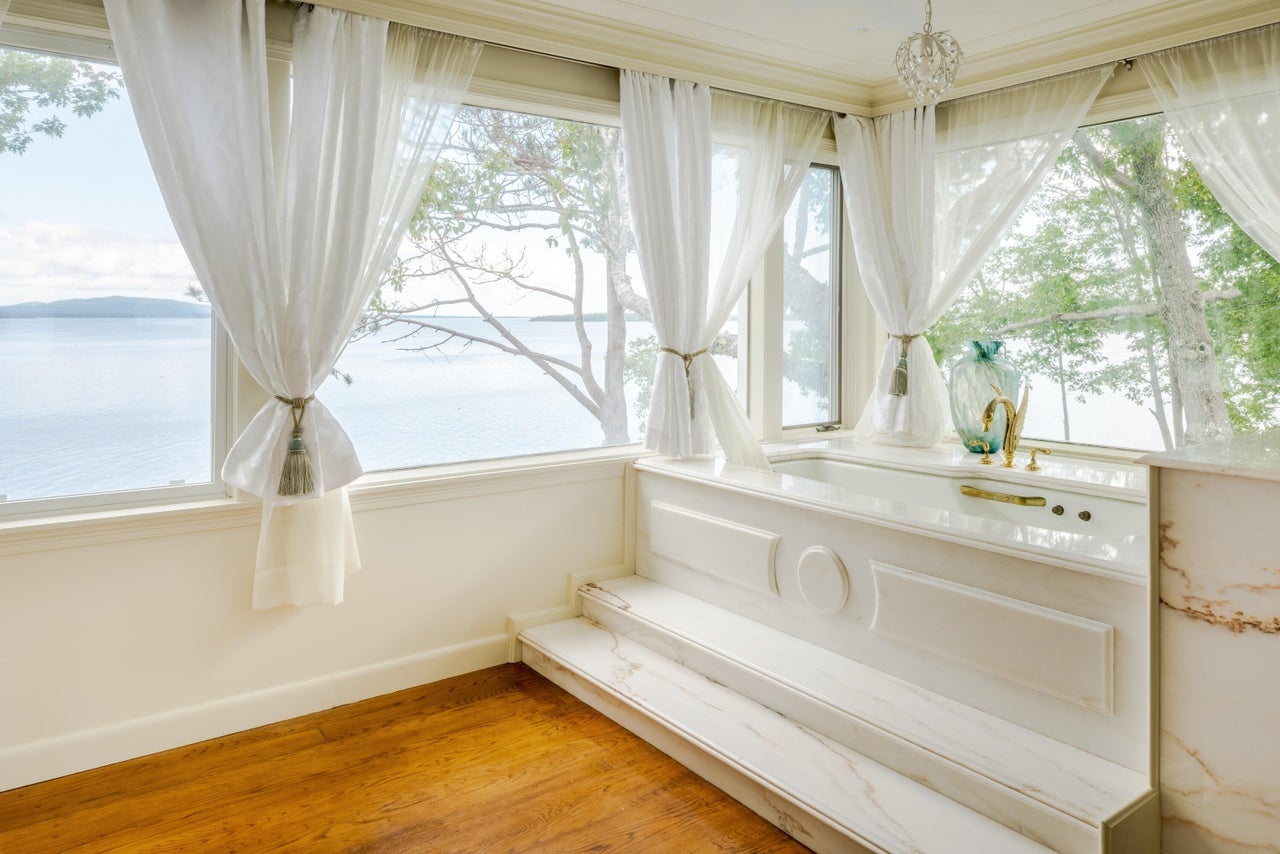A tub made of white stone sits in the corner of the room. Two small steps lead up to it. The rest of the visible floor is wood. Both walls are made up of picture windows that offer views of the ocean and distant mountains. There are sheer, white curtains tied up hanging from the walls.