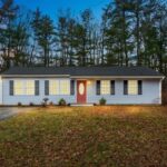 The exterior of a short ranch-style home at dusk. There are tall, evergreen trees in the background. the exterior of the home is light-blue, and the door is red.
