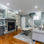 A greenish-gray living room with hardwood floors, white crown molding, a lighted gas fireplace with a stone surround, open shelving, a white-topped coffee table on black Y-shaped legs, and a light-gray couch.