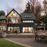 The exterior of this A-frame home is a dark gray color. The home appears to be two stories, with two balconies visible on the second floor. There are several huge, glass doors on both the first and second level, with lights on inside. The sun appears to be setting in the photo.