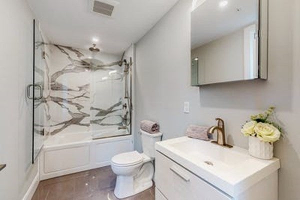 There is shower-tub combo with marble-patterned walls at the back of the room. The toilet sits in front of that, and a sink with white drawers underneath sits in front of that. A medicine cabinet mirror hangs about the sink.