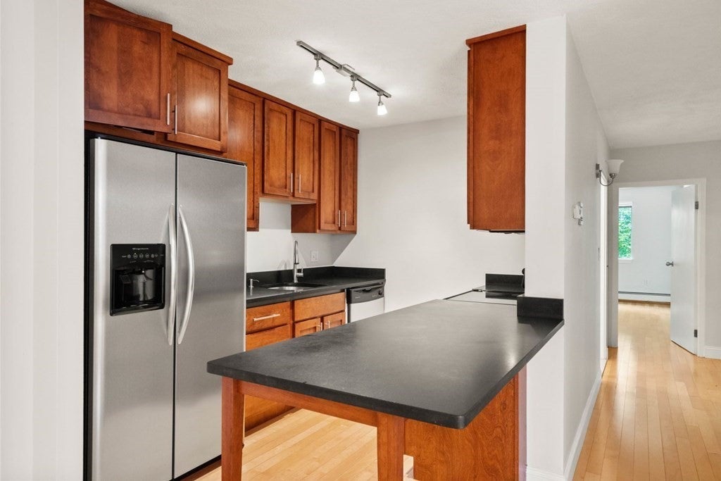 There is a stainless steal refrigerator and a row of wood cabinets along the back wall of this kitchen. A peninsula with a black countertop sit across from that. The room is brightly lit with a row of track lights, and the floor is wood. The hallway is visible on the left.