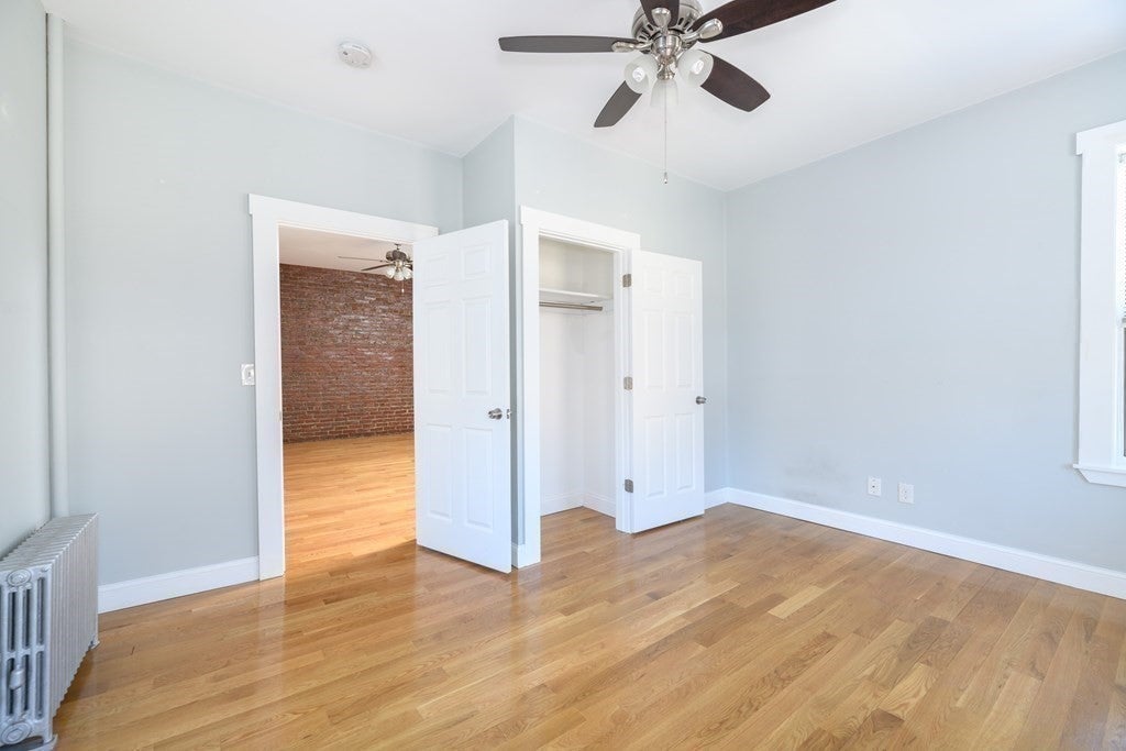 This room has wood floors and light gray walls. There's a ceiling fan hanging in the center of the room. There's a small closet to the right of the entrance and an exposed brick wall is visible in the the room over.