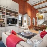 An L-shaped couch sits in the foreground of the this photo, facing a tall bookshelf with a TV and books. There is a circular, cushioned coffee table in front of the couch. The dining area and stairs up to the mezzanine are visible in the background on the right side.