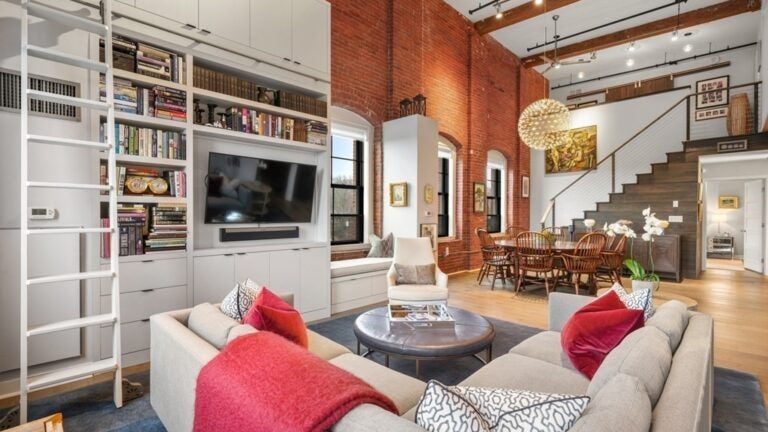 An L-shaped couch sits in the foreground of the this photo, facing a tall bookshelf with a TV and books. There is a circular, cushioned coffee table in front of the couch. The dining area and stairs up to the mezzanine are visible in the background on the right side.