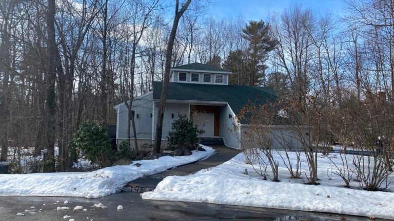 A view of the front facade, which features a prominent shed dormer with four square windows. A walkway curves slightly up to a sheltered, white front door. the sky is blue, the trees are bare, and the ground is covered in snow.