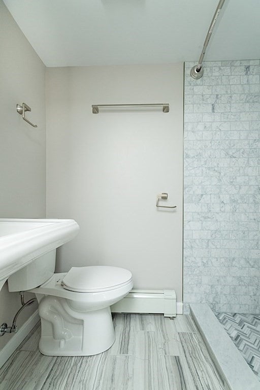 This bathroom has gray, wood-look floors and a gray tile walk-in shower. The room is small, and the toilet sits against the wall across from the shower. The white sink is just barely visible in the foreground.