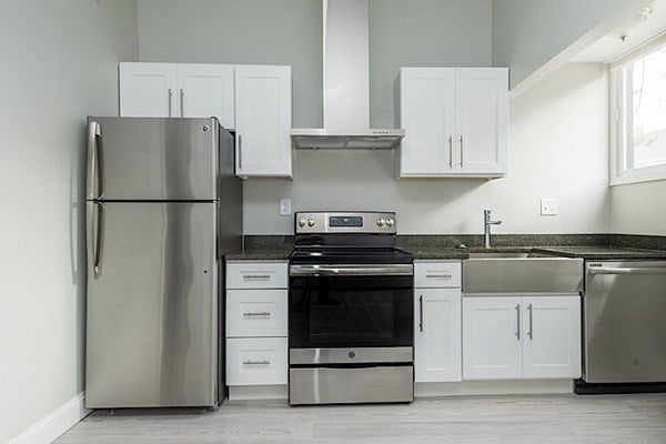 The room is all gray and white, with a stainless steal refrigerator on the far left, white cabinets overhead, and a stainless steal oven and dishwasher to the right of the fridge. There's also a stainless steal farmhouse sink to the left of the dishwasher. The floors are a gray, wood-look material.