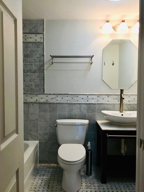 This view of the bathroom shows the toilet at the center of the room, which is lit with three sconce lights. These lights hang above a geometric mirror. A vessel sink sits under the mirror, atop a white counter and a dark cabinet. The walls and floors are decorative tile.