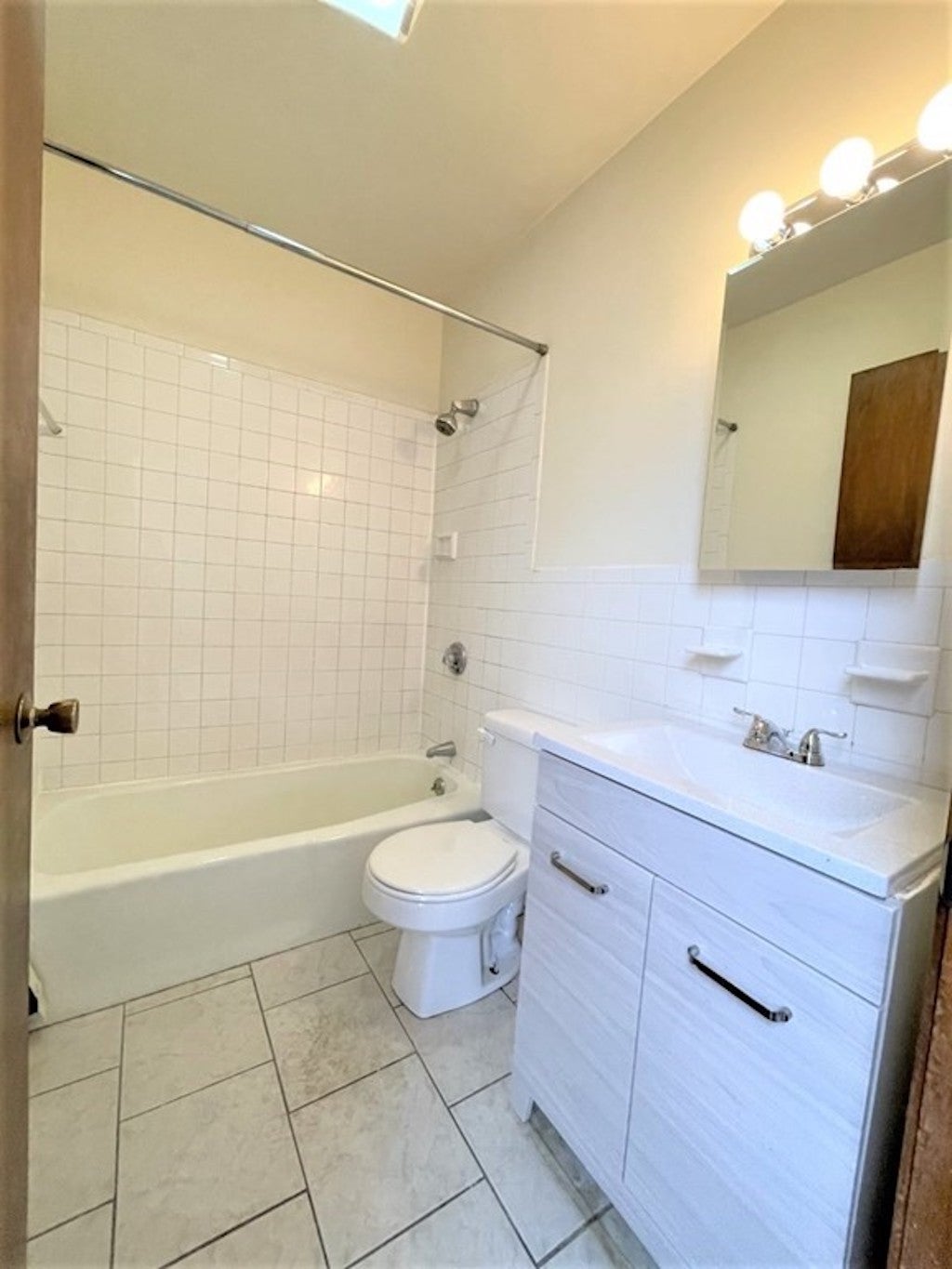The bathroom has a tile floor, white walls and a white ceiling. There's a single-sink vanity with white cabinets underneath. There's a small mirror with three spotlights overhead. The shower-tub combo takes up the space a the back of room.