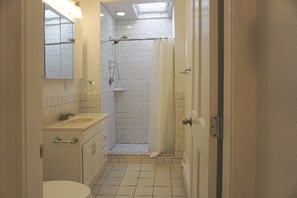 This bathroom is all white- white tile floors, white walls, and white counters. There's a subway tile shower in the back of the room, and white vanity with a sink along the left wall under a medicine cabinet mirror. The toilet is just visible in the left foreground.