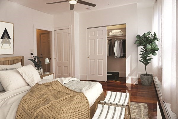 A queen size bed with a wooden headboard sits against left wall in the room. The floors are a dark, shiny wood, and the walls and ceiling are white. There's a ceiling fan with a light over the bed in the center of room, and two woven foot stool sit at the foot of the bed. There's a closet with a sliding door in the background. A lively house plant sits to the right of the closet.