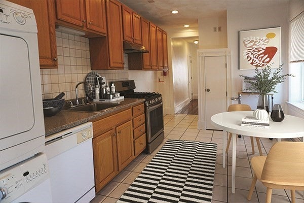 A stackable washer and dryer are visible in the left foreground of this photo of the kitchen. Next to this along the left wall there are wood cabinets hanging above the counter, which is a dark colored stone. There is a white dishwasher just to the right of the washer and dryer, and more cabinets to the right of that. The oven sits at the end of the counter. The floors are white tile, and there's a black and white striped sug between the counter and a small white table. Two wood chairs are set up at the table, which has a plant and a small of stack of book on it.