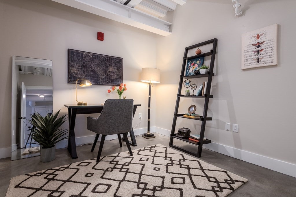 A modern bookshelf leans against the right wall of the room, which is lit with a lamp in the corner of the room. A cushioned chair is set up at a desk against the far wall. There is a patterned rug covering most of the floor in the room.