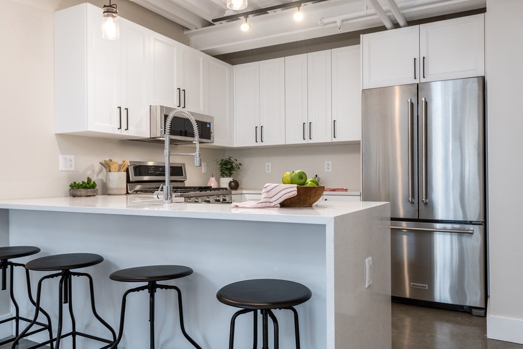 A double-door, stainless steal fridge sits in the right side of the kitchen. There are several wall-mounted, white cabinets along the wall, and there's a waterfall island in the foreground of the photo. There are four black, backless stools at the island.