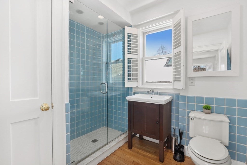 This bathroom has a walk-in shower with blue tile walls and a glass door in the left side. There is a window to the right of that, with white shutter, and a sink atop a dark wood vanity. The toilet is to the right of the sink. The same blue tiles line the bottom half of the walls outside the shower.