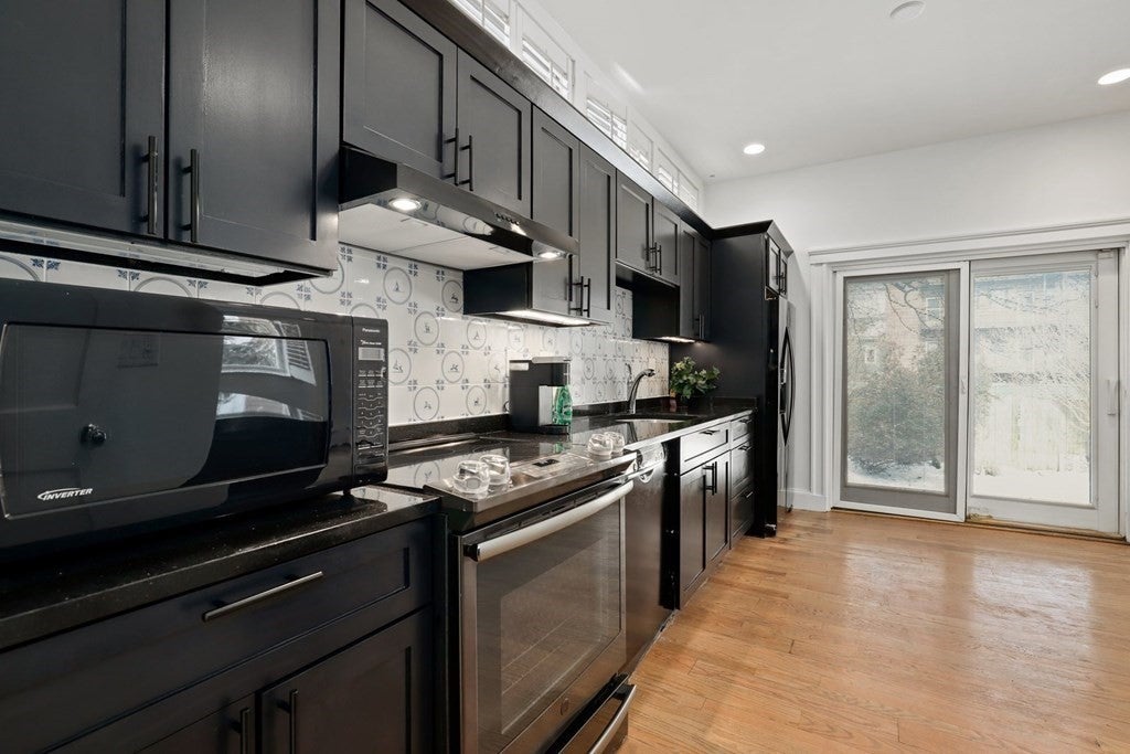 The kitchen has all black appliances and black cabinets along the left wall. There is a sliding glass door at the back of the room, and floors are wood. The space is well-lit with recessed lighting.