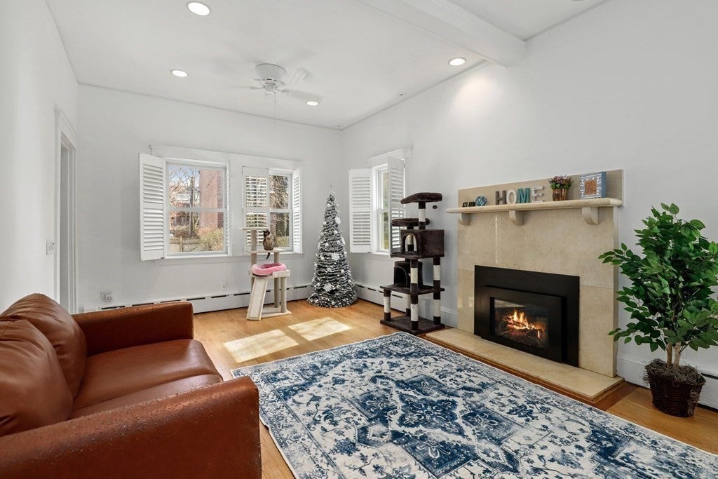 The living room has recessed lighting, white ceilings and white walls. There is a fireplace with a sand-colored tile surround in the right wall and a white and blue area rug on the floor in front of it. There is a brown leather love seat against the wall opposite the fireplace. In the background, there is a small Christmas tree in the corner and three windows letting in natural light.