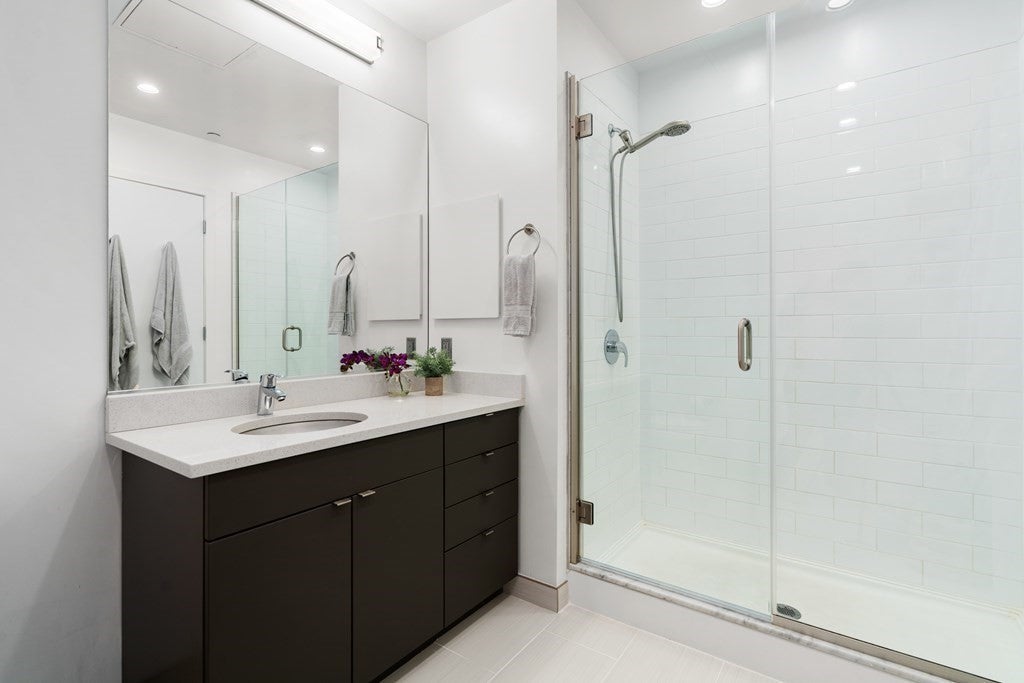 A vanity with a white countertop has a sink built in and dark wood drawers and cabinets underneath. The walk-in shower is to the right of the vanity, and has a detachable shower head and white, subway tile walls. There is a frameless mirror over the vanity.