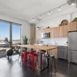 This spacious kitchen has shiny, black floors, white walls and a white ceiling. A track of spotlights hangs from the ceiling, and natural light enters through the sliding glass doors in the back wall. There is a tall table with a wood counter and four red stools and two grey stools set up around it. The stainless steal refrigerator is to the right of the room, and wood cabinets hang above and sit below the white counters. There is a peloton bike set up next to the sliding glass doors.