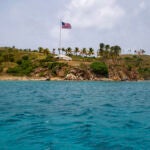 An ocean point view of an island with heavy vegetation, towering palm trees, and an American flag blowing in the breeze in a partly sunny day.