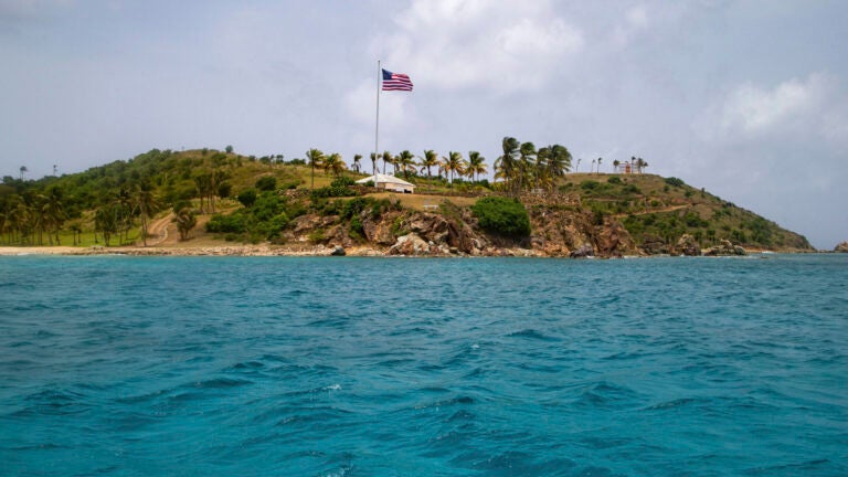 An ocean point view of an island with heavy vegetation, towering palm trees, and an American flag blowing in the breeze in a partly sunny day.