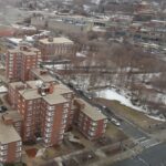 An aerial view of a snow-dotted landscape with multi-story residential buildings to the left and an empty lot with trees to the right.