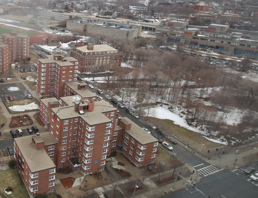 An aerial view of a snow-dotted landscape with multi-story residential buildings to the left and an empty lot with trees to the right.