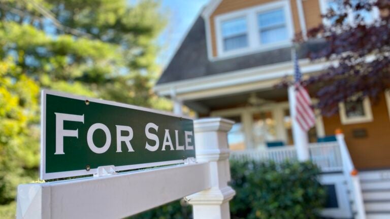 A for sale sign sits in front of a pumpkin-colored home with a farmer's porch and green shrubs and trees.