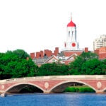 John W. Weeks Bridge and clock tower over Charles River in Harvard University campus in Boston with trees and blue sky.